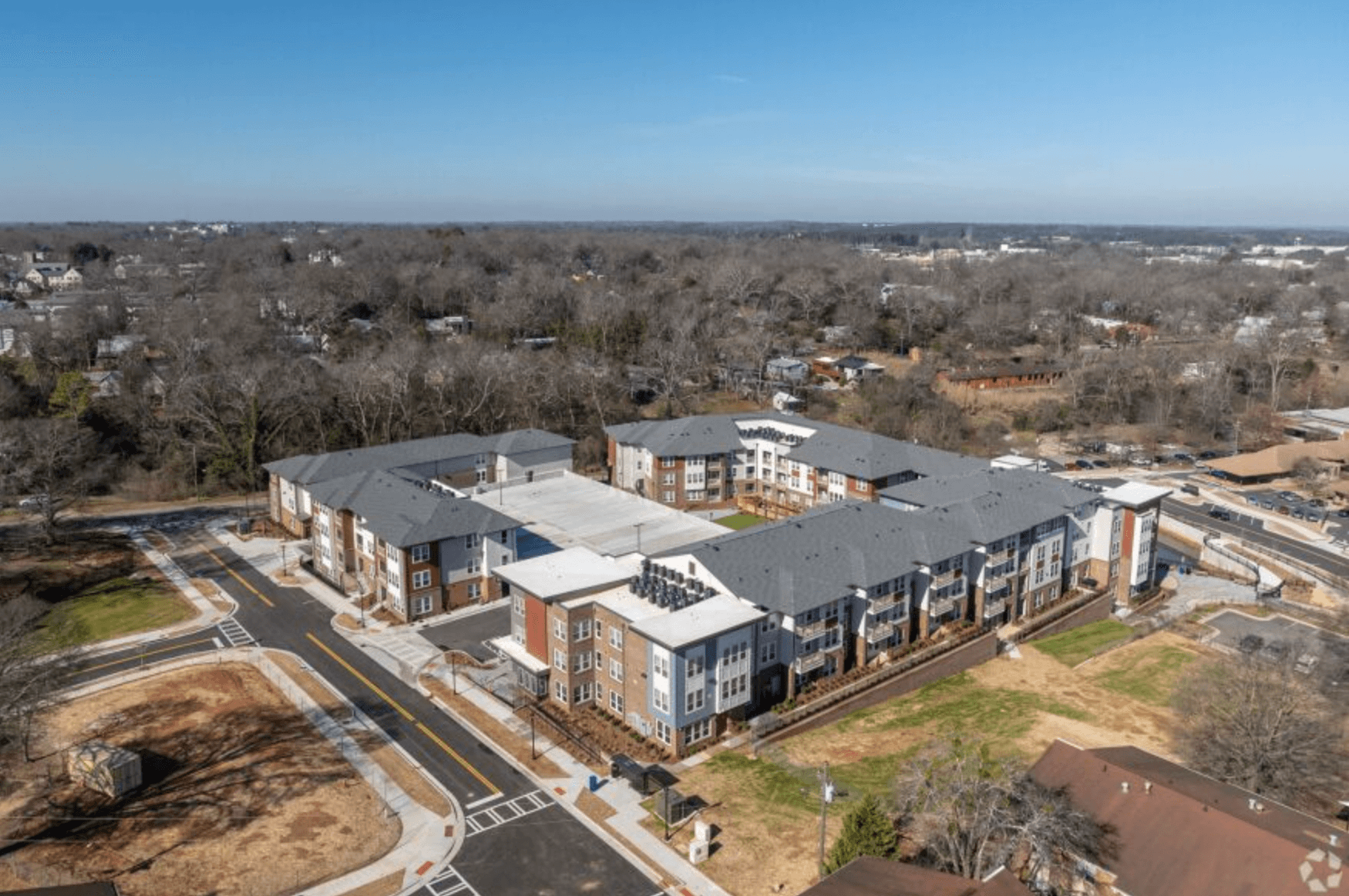 Aerial view of The View at NoDA with multiple three-story buildings, featuring a mix of brick and white panel exteriors, pitched gray roofs, and landscaped walkways. The development is surrounded by roads, green spaces, and a wooded area in the background.